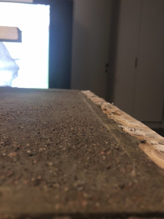 En betonskive med ufuldkommen overflade, med en træliste langs kanten i et indendørs miljø. Skiven virker en smule tyndere end ønsket.