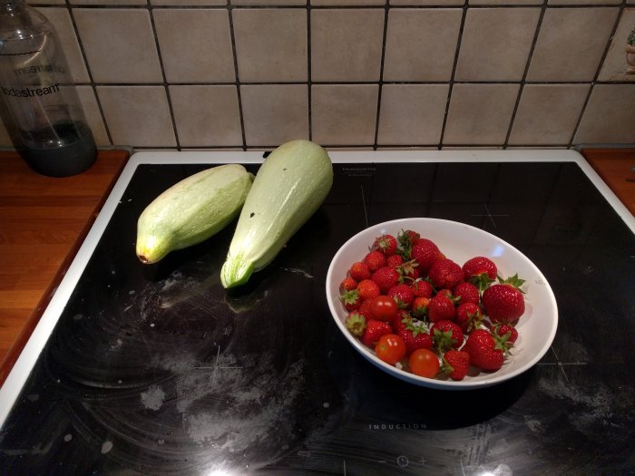 Två stora gröna squash och en skål med röda jordgubbar och tomater på en spishäll.