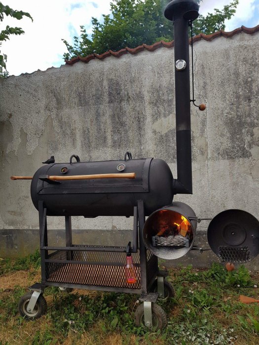 Svart rökare/grill på hjul med brinnande träkol, närbild av eldstaden, mot en grå vägg.