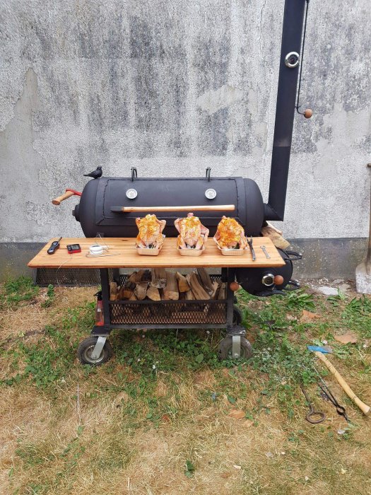 Svart rökgrill med öppet lock och fyra kycklingar på spett framför en grå vägg, trädgårdsredskap på marken.