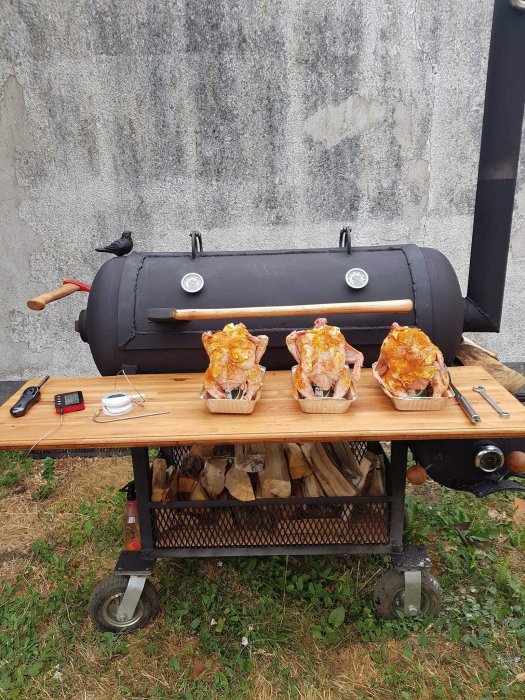 Rökgrill utomhus med tre kycklingar på spett och grillverktyg på arbetsbänk.