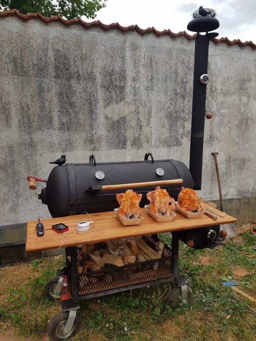 Svart rökgrill på hjul med två kycklingar på sidobordet, grillverktyg och ved förvarad undertill framför grå betongvägg.