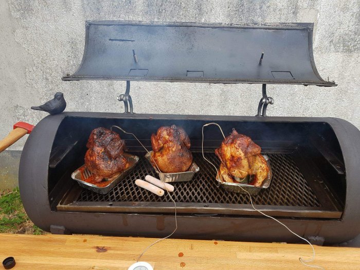 Tre grillade kycklingar i en stor öppen rökgrill utomhus med en fågel som sitter på dess kant.