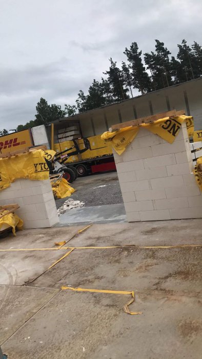 Ny leverans av byggmaterial med balkar och ublock vid en byggarbetsplats, skyddade av DHL-presenningar.