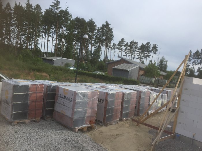 Byggplats med levererade byggblock och material, blivande hus vägg och skog i bakgrunden.