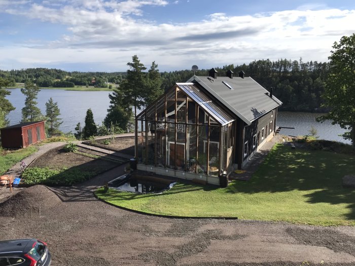 Bild av ett modernt hus med stora glaspartier och mörkt tak vid en sjö med naturen och landskapet synligt i bakgrunden.