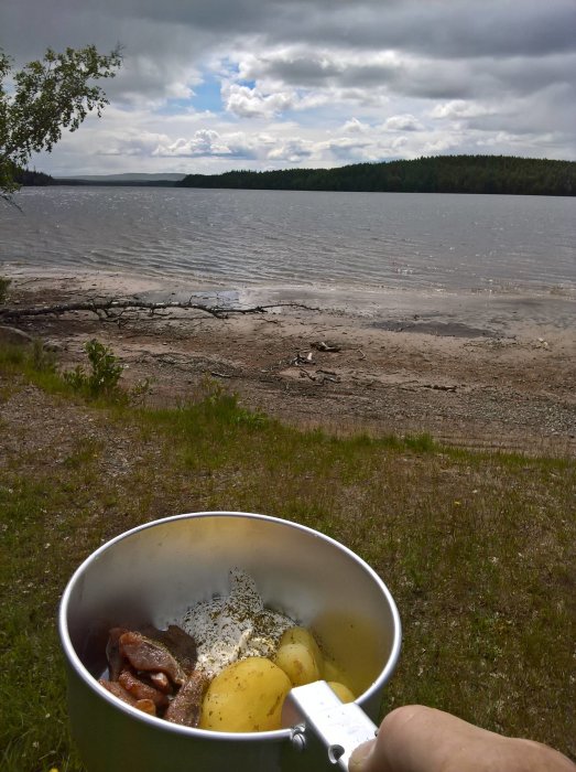 En gryta med potatis och kött som hålls av en person, med en sjö och skog i bakgrunden under midsommarfirandet i Dalarna.