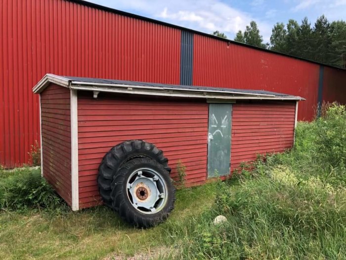 Gammal arbetsbod i rött med traktordäck vid sidan, omgiven av högt gräs, med en stor lada i bakgrunden.