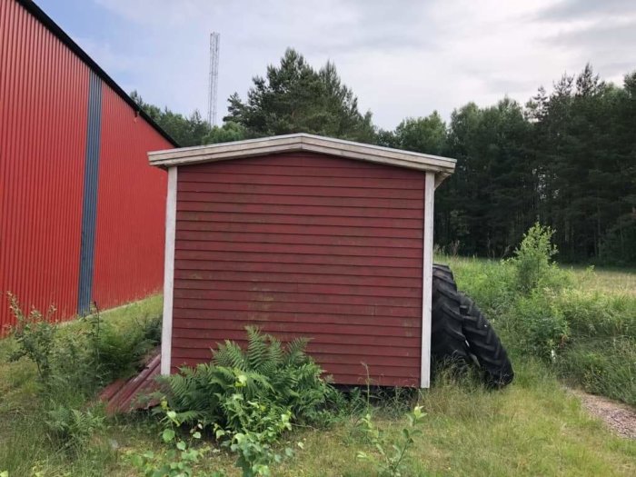 Röd träarbetsbod står i grönskande mark med en stor däckstapel vid sidan och en röd byggnad i bakgrunden.