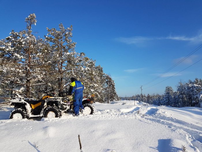 Person i blå overall bredvid en fyrhjuling i ett snötäckt landskap med träd under blå himmel.