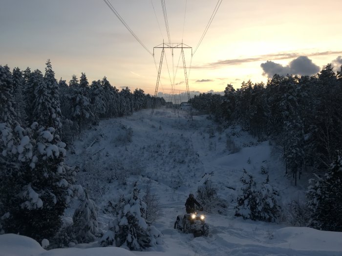 Vinterlandskap med snötäckta träd och terrängfordon under en dramatisk solnedgång.