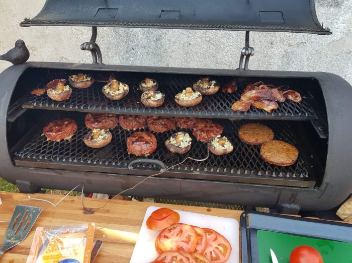 Hamburgare och fyllda champinjoner på en rökgrill med skivade tomater på skärbräda.