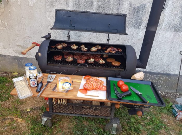 Grill med öppet lock fullt av hamburgare, ett skärbräde med skivade tomater och burgartillbehör vid sidan.