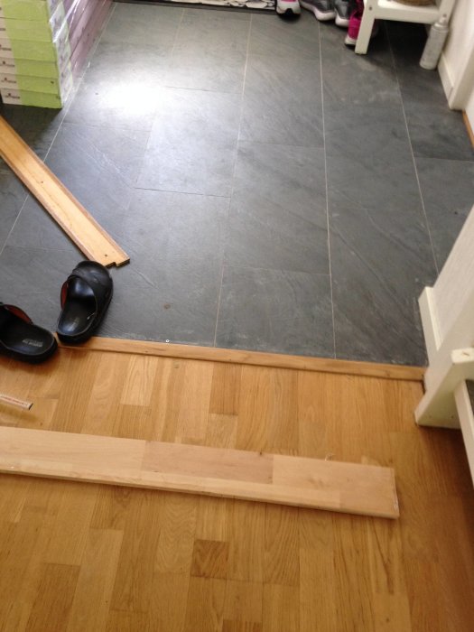 Trägolv i övergång till klinkers, med synlig höjdskillnad och ett löst trästycke på golvet.
