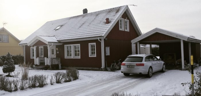 En snöklädd röd svensk villa med vit snickarglädje och carport med en bil parkerad under.