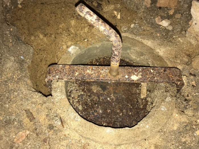 Rostig lucka i källargolv med handtag, troligen renslucka för avlopp, inramad av betong och grus.