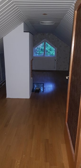 Övervåning före renovering med snedtak, golv av trä och tapetserade väggar.