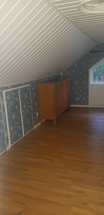 Övervåning före renovering med snedtak, blommig tapet och trägolv, möbel syns.