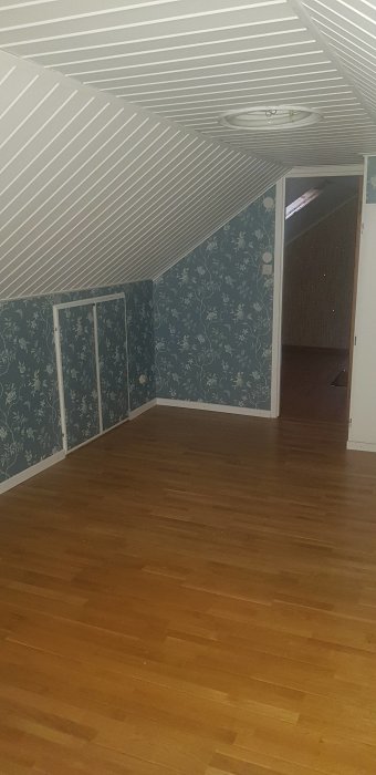 Övervåning före renovering med snedtak, parkettgolv och vägg med blommigt tapetmönster.