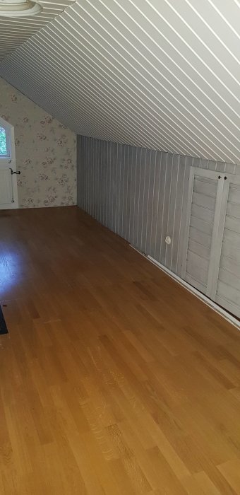 Inredd övervåning före renovering med snedtak, vita strimmiga väggar och blommigt tapetserad vägg.