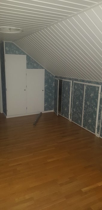 Övervåning före renovering med snedtak, parkettgolv och tapetserade väggar.