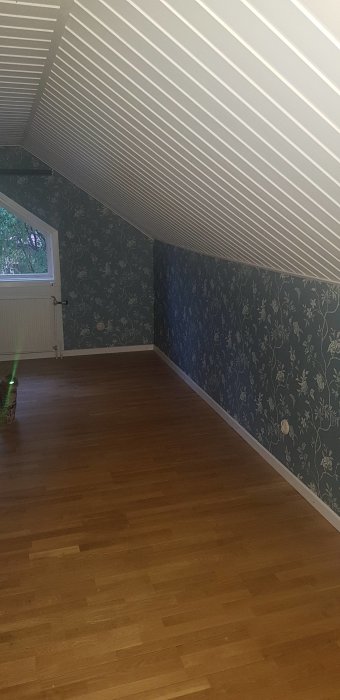 Övervåning före renovering med snedtak, blommiga tapeter och trägolv.