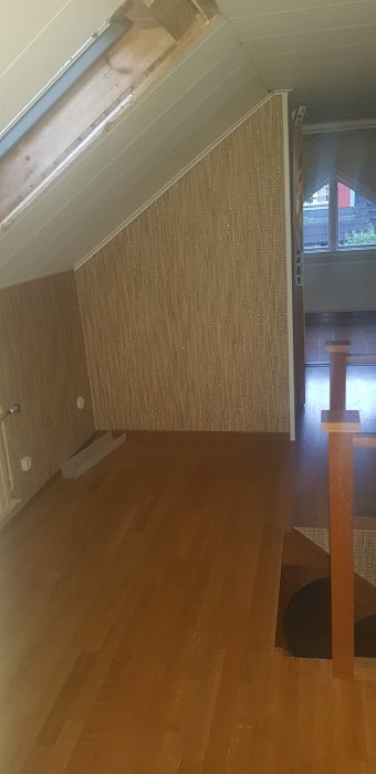 Övervåning före renovering med snedtak, brun tapet, trägolv och öppet takskott.