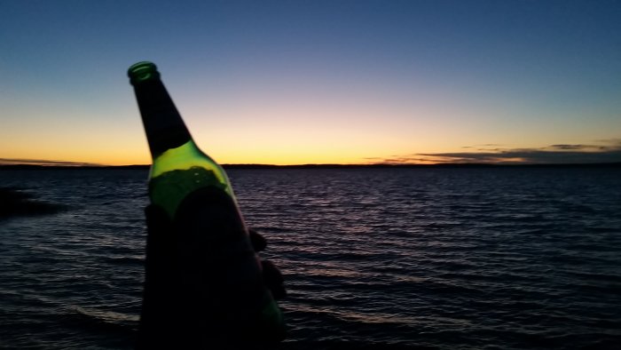 Solnedgång över havet på midsommarnatten med en ölflaska i förgrunden.