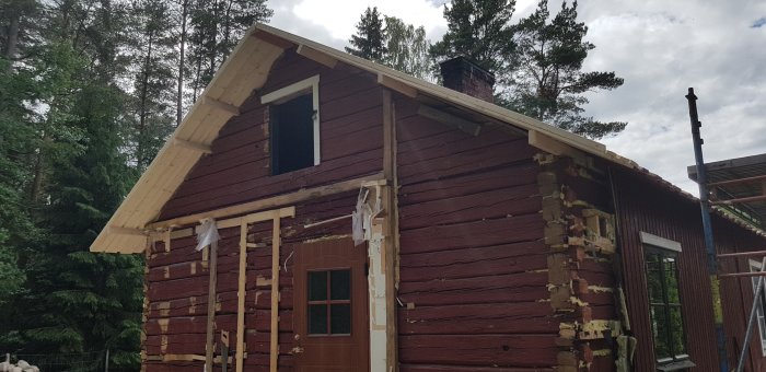 Renovering av rött trähus där fasaden delvis är avtäckt och nytt takstolsarbete pågår, omgivet av skog.