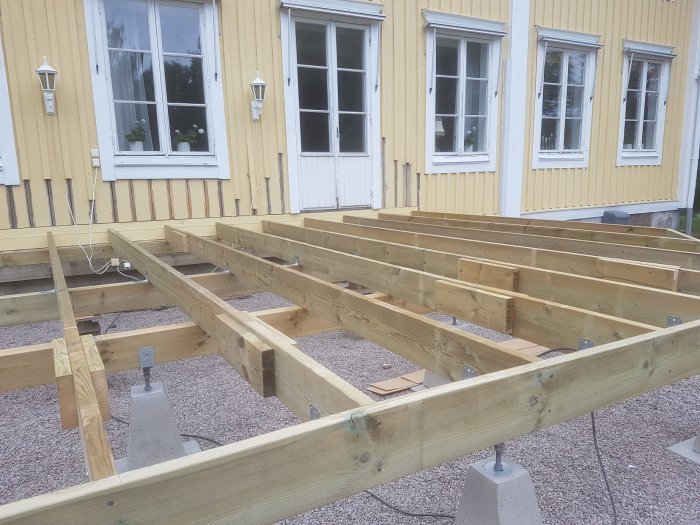 Nybyggda träreglar på ett byggprojekt framför ett gult hus med vita fönster.