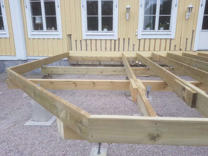 Altanens trästomme med fasade ytterhörn i 45 grader, bredvid ett gult hus.