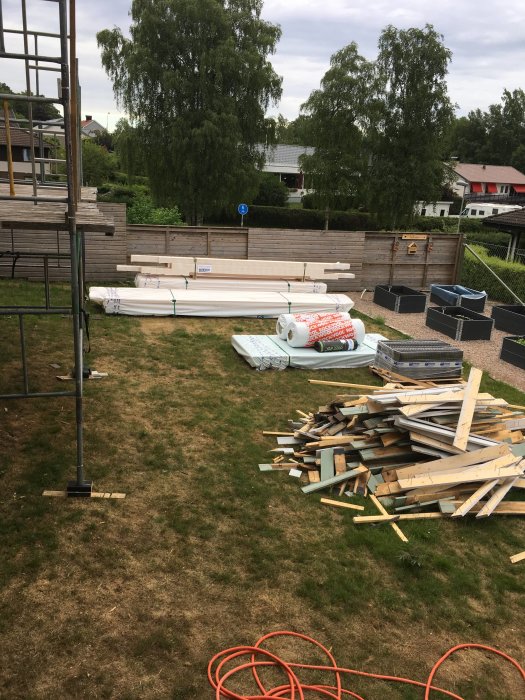 Byggmaterial och verktyg ordnade på gräsmatta inför byggprojekt med byggnadsställning i bakgrunden.