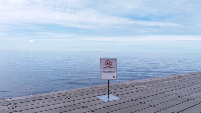 Träbrygga i Malmö med skylt som indikerar badförbud, mot bakgrund av lugnt hav och blå himmel.