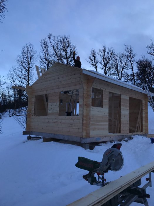 Person i arbetskläder på taket av en nybyggd attefallare i skymningen, med snötäckt mark och trädstammar i bakgrunden.