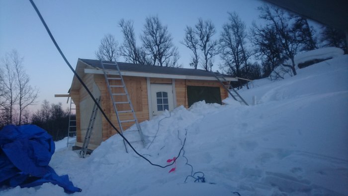 En halvfärdig byggnad i timmer under kvällshimmel, omgiven av snö, med stegar lutade mot fasaden och en täckpresenning till vänster.