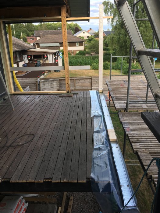 Pågående arbete på en trädäckskonstruktion med delvis lagda träplankor och utsikt mot grannhus.