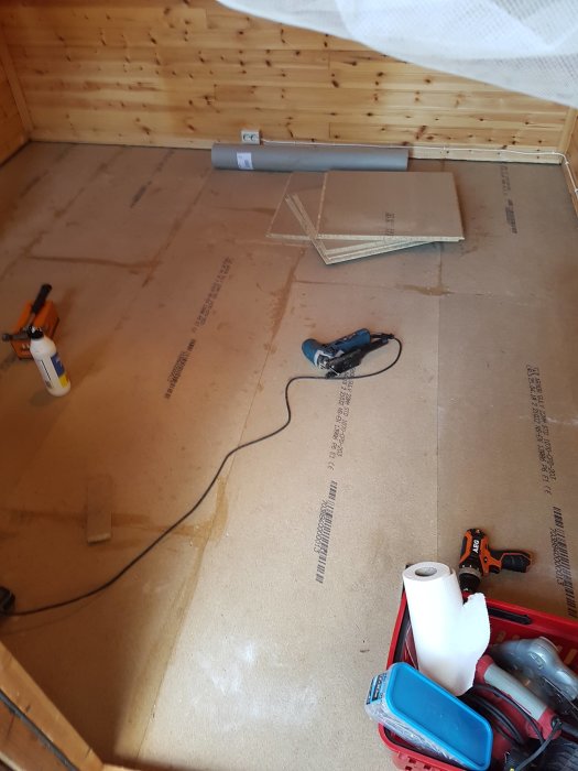 Renovering pågår med verktyg och byggmaterial på golv med skyddspapp i ett rum med träpanel.