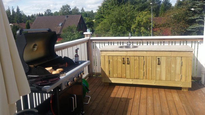 Hembyggt utekök med betongskiva och trästomme, utrustat med diskho, på en träaltan bredvid en grill.