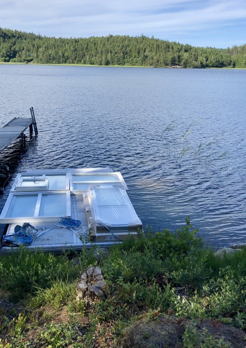 Flotte med fönster och dörrar på en sjö nära en brygga, med skog i bakgrunden.