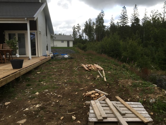 Ojämnt utspridda jordmassor nära ett hus med träterrass och skog i bakgrunden, sten och byggmaterial framför.