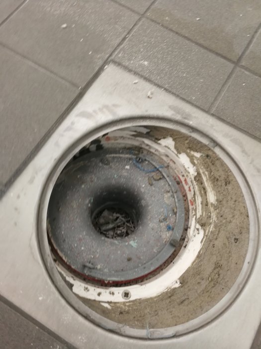 Golvbrunn under konstruktion med skräp, plast och murbruk i avloppet och vattenlåset under klinkerramen.