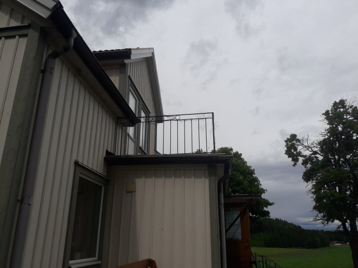 Balkong med järnräcke på ett tvåvåningshus, diskussion om byte till trä, under molnig himmel.