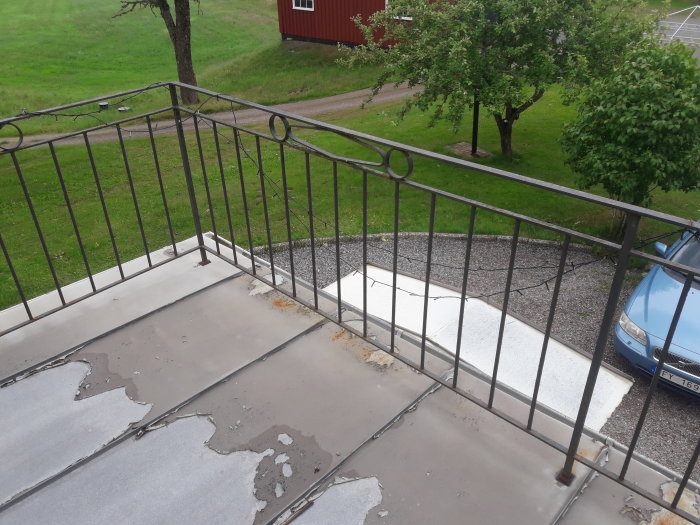 Äldre järnräcke på en balkong med plåtgolv och utsikt över en trädgård och parkerad bil.