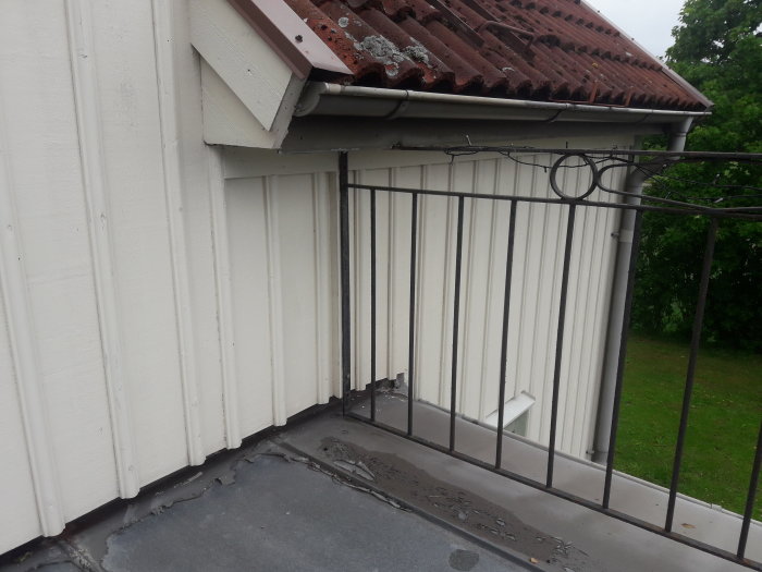 Järnräcke på en balkong med vertikala spjälor och två ringar inuti, monterat mot en vit husvägg och ett plåtgolv.