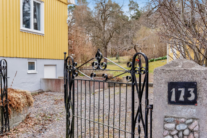 En ståtlig, smidesjärnsgrind framför en gul husfasad med numret 173, omgiven av en oanlagd tomt och lövträd.