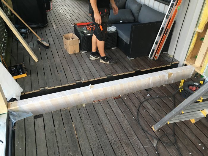 Delvis demonterad trätrall med verktyg och byggmaterial, samt en person som syns delvis, på en balkong under renovering.