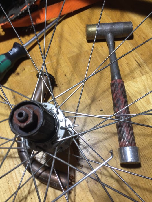 Cykelhjul med nav och ekrar på ett trägolv, nära en hammare och ett improviserat verktyg av kopparrör.