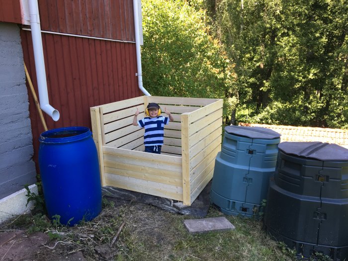 Ett barn står i en nybyggd träkompostlåda bredvid gamla matkomposttunnor utomhus.