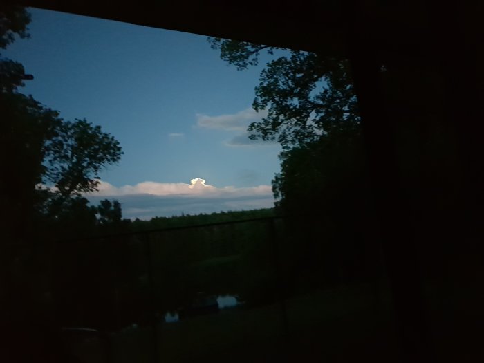 Nattvy genom ett fönster med träd, räcke och ett delvis molntäckt månsken.