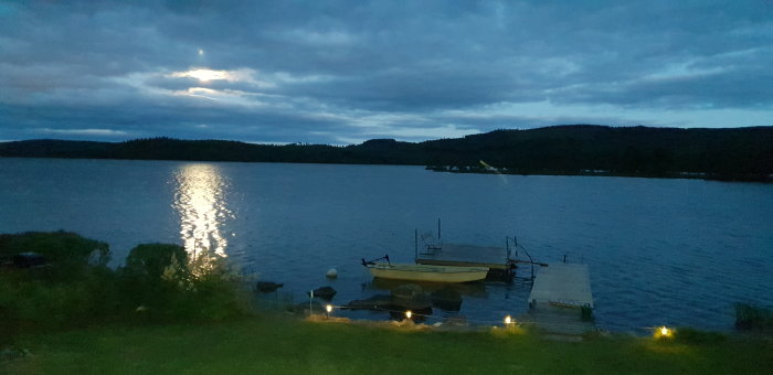 Kvällsbild av en sjö med månens spegling på vattenytan, båt vid en brygga och ljus på stranden.